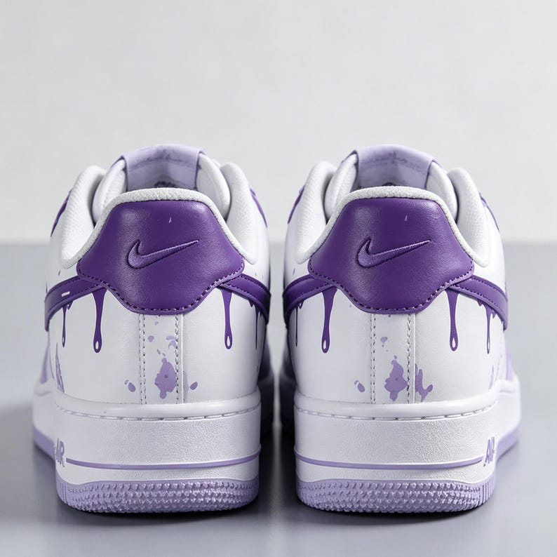 May include: Pair of white and purple sneakers with a low-top design. The sneakers feature a purple heel tab with a Nike logo and purple paint drip details. The soles and accents are also purple.