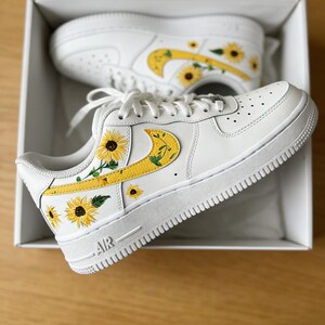 May include: White sneakers with yellow sunflower designs. The shoes feature a white sole, laces, and a yellow swoosh with a sunflower and green leaf design. The sunflower design is repeated on the side and toe of the shoe.