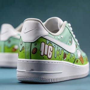 May include: Custom-painted sneakers with a green and white color scheme. The shoes feature a white Nike swoosh and sole, with a design that includes the word "JIG" and illustrations of coffee beans and ice cream cones. The background is a light green.