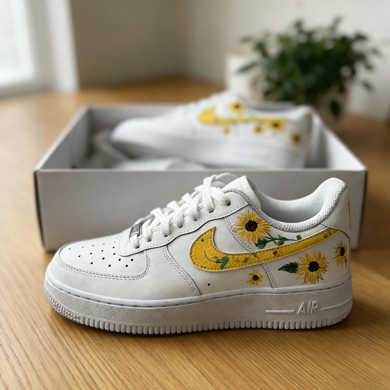 May include: White leather sneakers with yellow sunflower embroidery. The sneakers feature a yellow swoosh and sunflower details on the side and heel. The shoes are in a white box, with another pair visible.
