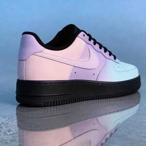 Op de afbeelding: Een pastelkleurige sneaker met een zwarte zool. De schoen heeft een gradi&euml;ntontwerp, dat overgaat van roze naar paars naar lichtblauw. De iconische Nike swoosh is zichtbaar aan de zijkant. De schoen wordt weerspiegeld in het oppervlak eronder.