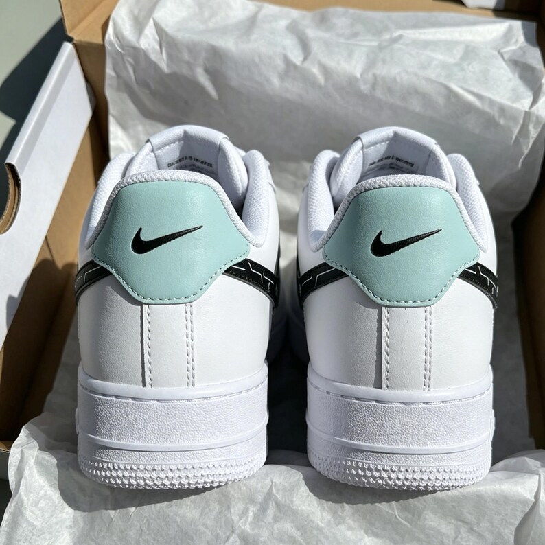 May include: Pair of white sneakers with a light blue heel tab and black swoosh logo. The shoes have a white sole and are in a cardboard box.