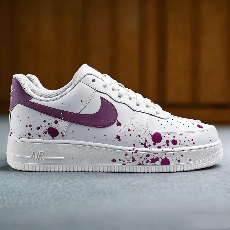 Puede incluir: Zapatilla de cuero blanca con un logotipo morado y dise&ntilde;o de salpicaduras de pintura morada. La zapatilla tiene una suela blanca con "AIR" impreso en el lateral. Una zapatilla deportiva elegante y moderna.