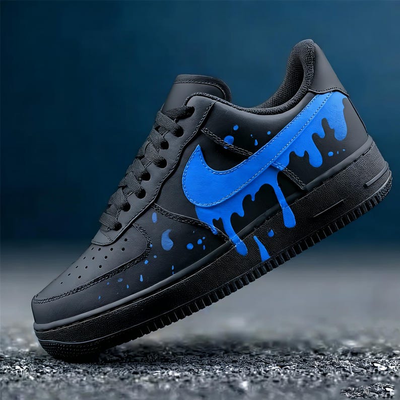 May include: Black and blue Nike Air Force 1 sneaker. The shoe features a black base with a blue Nike swoosh and blue paint splatter design. The sneaker has a black sole and laces.