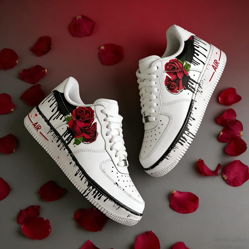 May include: White sneakers with red rose designs and black paint drip accents. The shoes have the word "AIR" in red on the side. Red rose petals are scattered around the shoes.