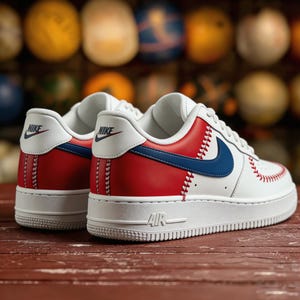 Op de afbeelding: Witte en rode sneakers met blauwe accenten, met honkbal-ge&iuml;nspireerde stiksels en een blauwe Nike swoosh. De sneakers hebben een witte zool met "AIR" aan de zijkant. De achtergrond is wazig, met kleurrijke bollen.