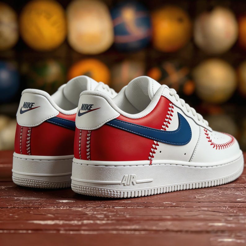 Op de afbeelding: Witte en rode sneakers met blauwe accenten, met honkbal-ge&iuml;nspireerde stiksels en een blauwe Nike swoosh. De sneakers hebben een witte zool met "AIR" aan de zijkant. De achtergrond is wazig, met kleurrijke bollen.