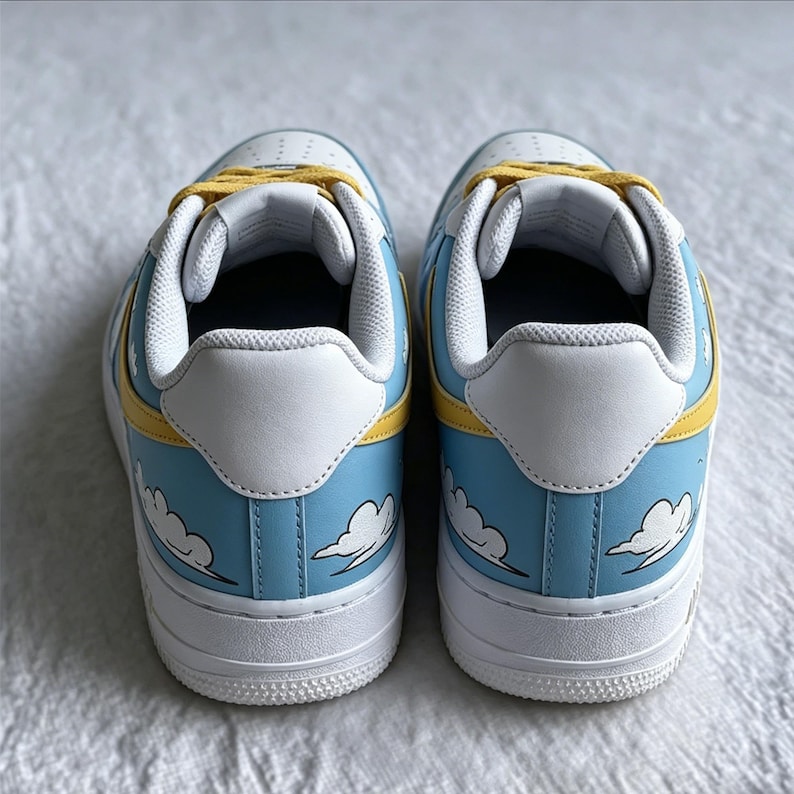 May include: Pair of white trainers with a light blue and yellow design. The trainers feature a cloud pattern on the sides, yellow laces, and white soles. The shoes are designed for casual wear.