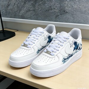 May include: Pair of white sneakers with a blue wave design on the side. The shoes have white laces, a white sole, and the Nike swoosh logo. The design is inspired by the famous Japanese artwork "The Great Wave off Kanagawa."