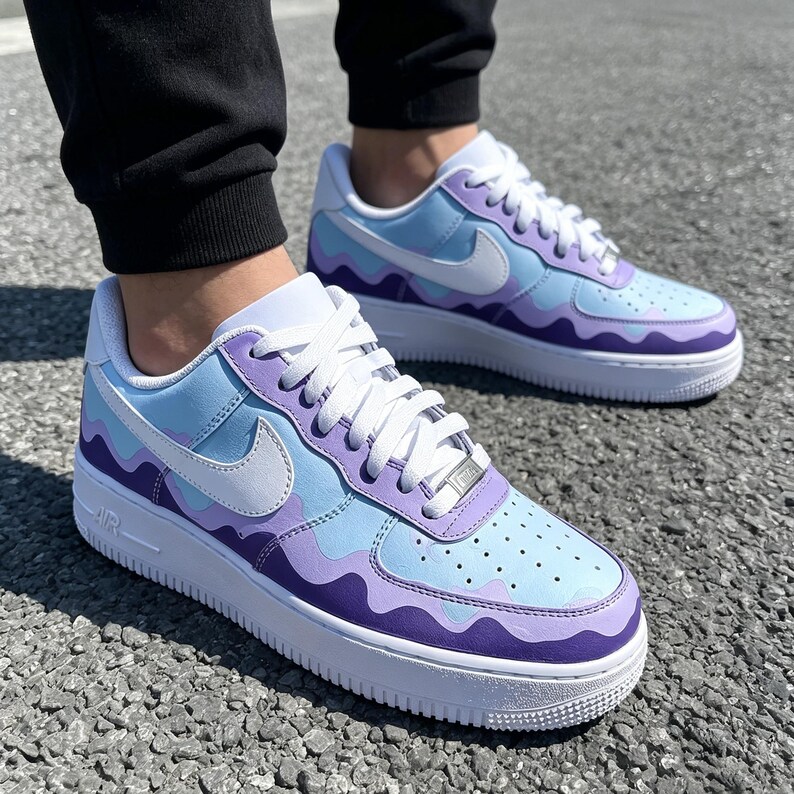 May include: A pair of custom-painted Nike Air Force 1 sneakers. The shoes feature a white sole, white laces, and a light blue, lavender, and purple wavy design. The Nike swoosh is white.