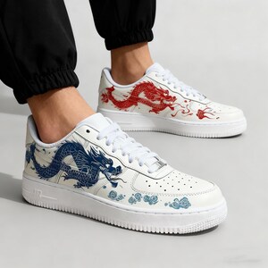 May include: White sneakers with a low-top design, featuring a dragon graphic. One shoe has a red dragon, the other a blue dragon. The shoes have white laces and soles, with a perforated toe box.
