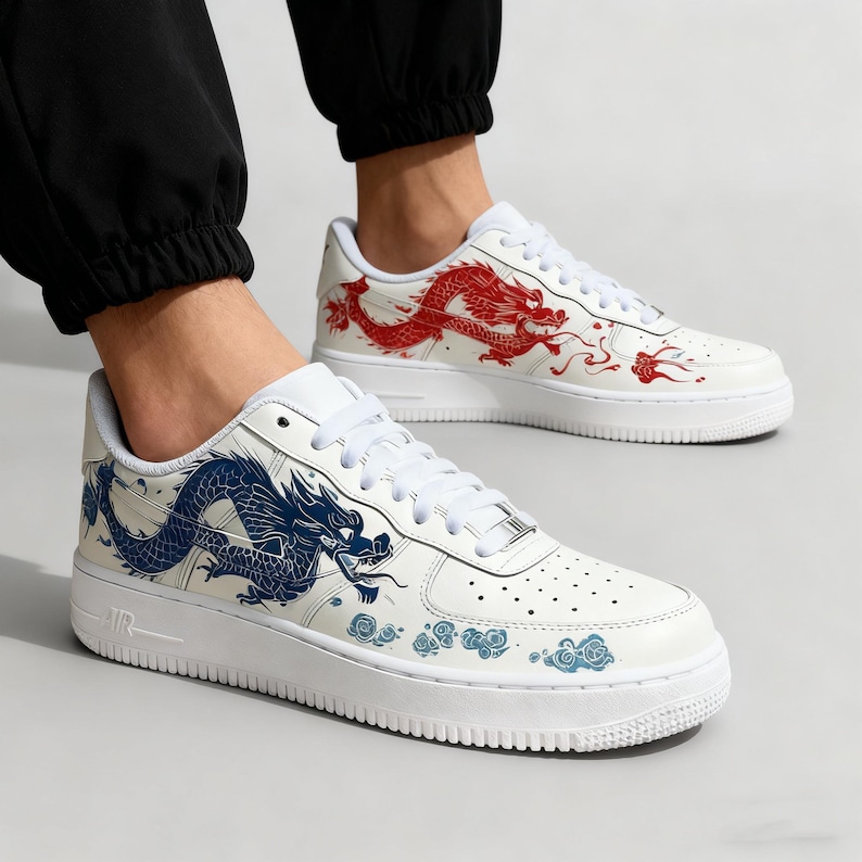 May include: White sneakers with a low-top design, featuring a dragon graphic. One shoe has a red dragon, the other a blue dragon. The shoes have white laces and soles, with a perforated toe box.