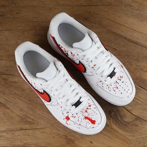 Może przedstawiać: Białe trampki z czerwonymi i czarnymi rozpryskami farby. Buty mają czerwone logo swoosh i białe sznurowadła. Trampki są w klasycznym sportowym stylu.