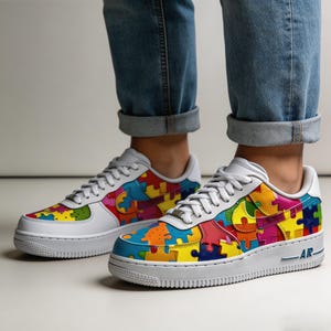 May include: White sneakers with colorful puzzle piece design. The shoes have a white sole and laces, with the iconic swoosh logo in a puzzle piece pattern. The shoes are worn with blue jeans.