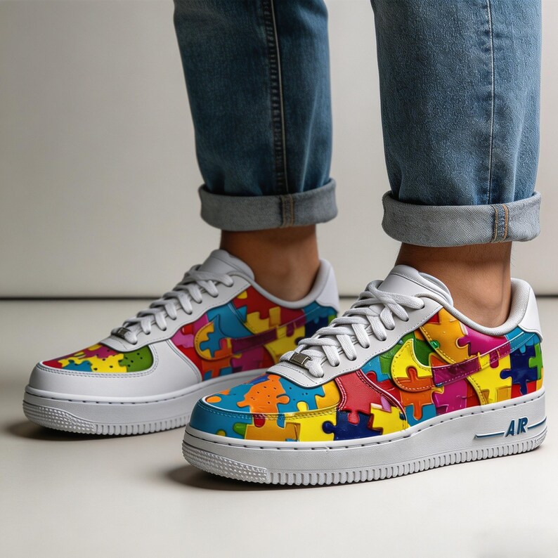 May include: White sneakers with colorful puzzle piece design. The shoes have a white sole and laces, with the iconic swoosh logo in a puzzle piece pattern. The shoes are worn with blue jeans.