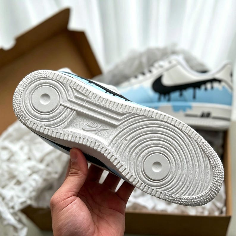 Puede incluir: Zapatillas Air Force 1 blancas y celeste con detalles negros. Las zapatillas presentan un dise&ntilde;o &uacute;nico con un efecto de pintura goteando. La suela es blanca con un patr&oacute;n circular y el logo de Nike.