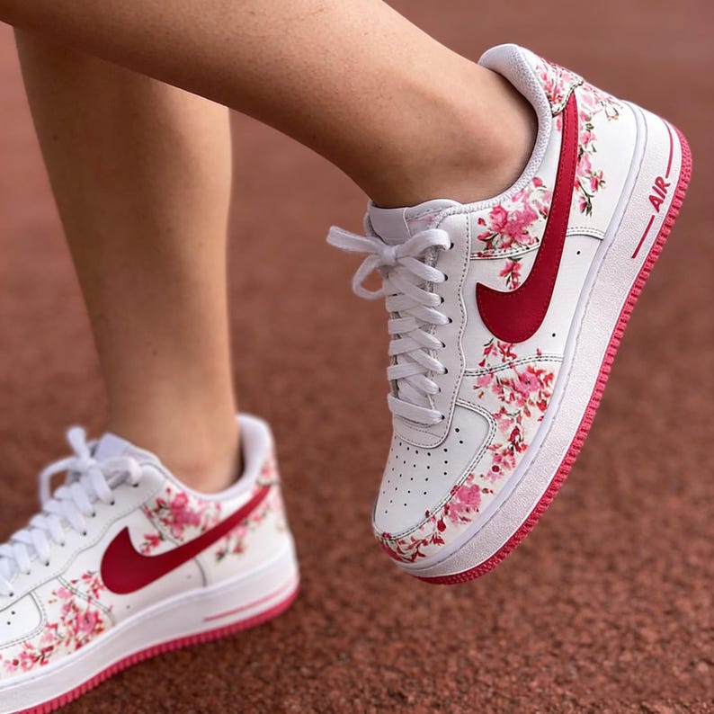 May include: White trainers with a floral pattern in pink and red. The trainers feature a red swoosh and a red sole. The word "AIR" is printed in red on the side.