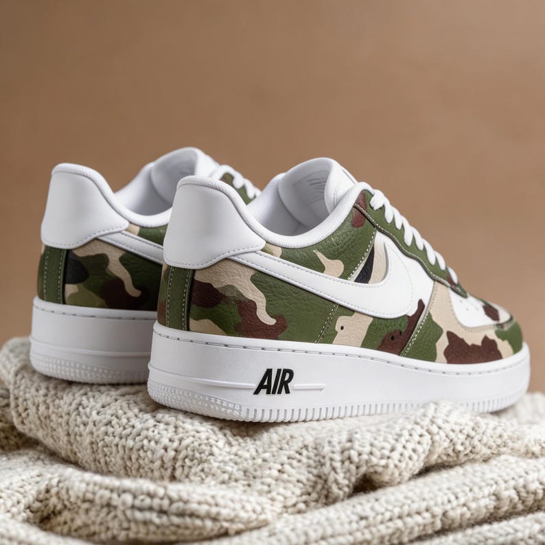 May include: A pair of white-soled sneakers with a camouflage pattern in shades of green, brown, and tan. The sneakers have white accents, including the iconic swoosh and heel tab. The word "AIR" is printed in black on the side of the white sole.