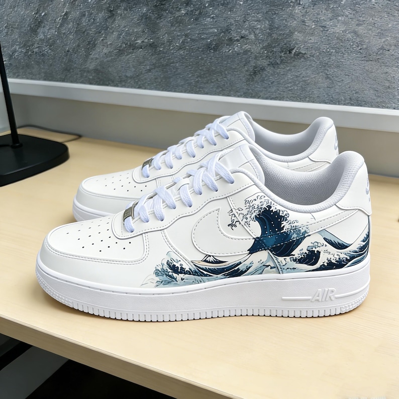 May include: White sneakers with a wave design in shades of blue and gray. The shoes have white laces, a white sole, and the word "AIR" on the side. The design is inspired by Japanese art.