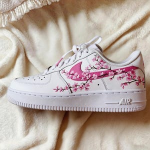 May include: White athletic shoe with a pink floral design. The shoe features a pink swoosh and pink cherry blossom branches. The word "AIR" is visible on the side of the white sole.