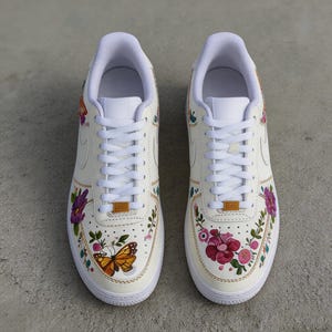 May include: Pair of cream-colored sneakers with floral and butterfly designs. The shoes feature white laces, soles, and inner linings. The painted flowers are in shades of pink, purple, and red, with a yellow and brown butterfly.