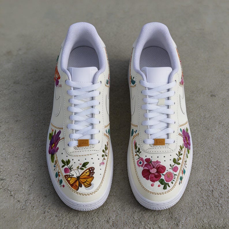 May include: Pair of cream-colored sneakers with floral and butterfly designs. The shoes feature white laces, soles, and inner linings. The painted flowers are in shades of pink, purple, and red, with a yellow and brown butterfly.
