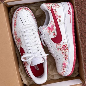 May include: White trainers featuring a pink and red floral design. They have a red swoosh and trim, with the word "AIR" in red. The trainers are presented in a cardboard box.