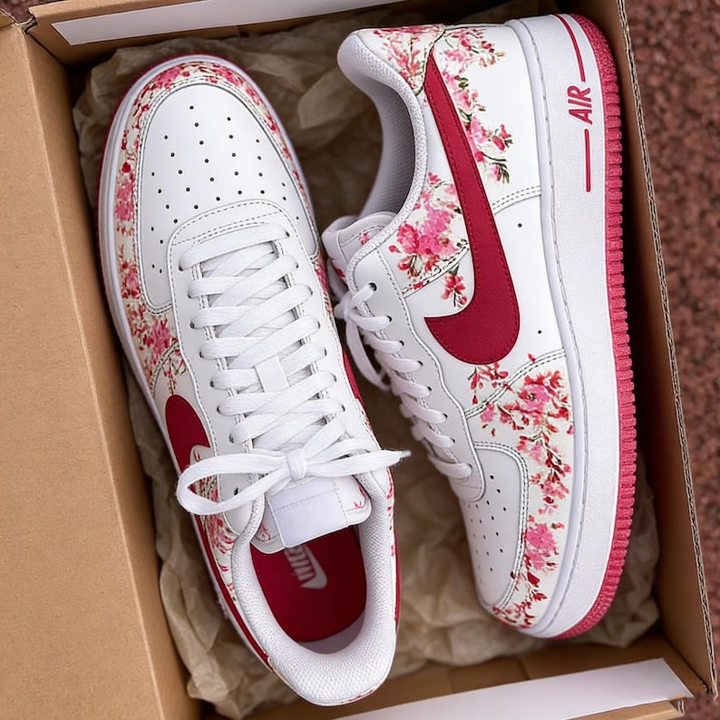 May include: White trainers featuring a pink and red floral design. They have a red swoosh and trim, with the word "AIR" in red. The trainers are presented in a cardboard box.