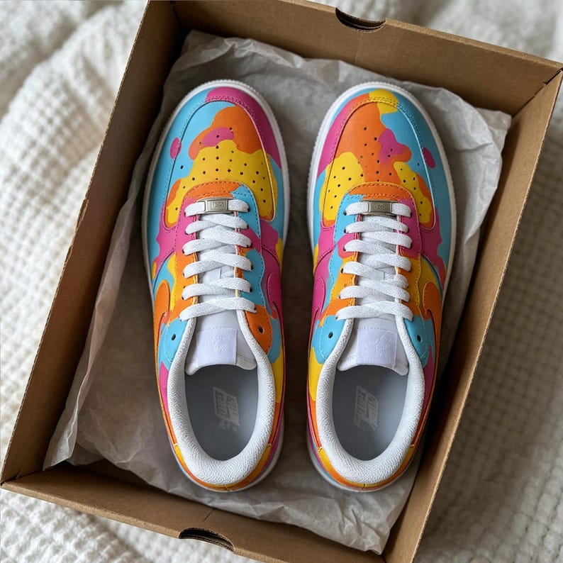 May include: A pair of colorful sneakers with a vibrant camouflage pattern in a cardboard box. The shoes feature a mix of pink, orange, yellow, and blue hues, with white laces and soles. The sneakers are new and unworn.