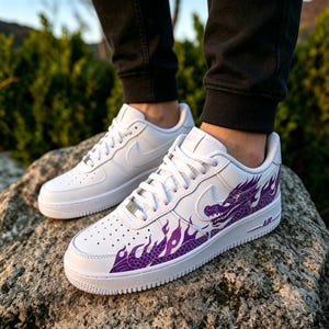 May include: White leather sneakers with a purple dragon design on the side. The sneakers have a classic low-top design with a white sole and laces. The Nike swoosh is visible on the side. The word "AIR" is printed on the side.