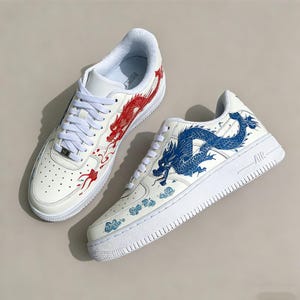 May include: Pair of white sneakers with red and blue dragon designs. The sneakers have white laces, soles, and a classic low-top design. The dragon artwork is detailed and adds a unique, artistic flair to the footwear.