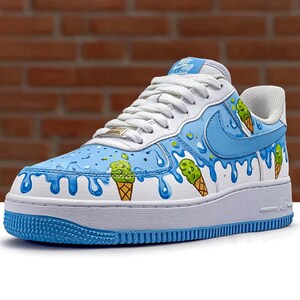Puede incluir: Zapatilla blanca y azul con dise&ntilde;o de cono de helado. El zapato presenta una base blanca con detalles azules, incluyendo un efecto de pintura goteando y gr&aacute;ficos de conos de helado. La suela es blanca con ribete azul.
