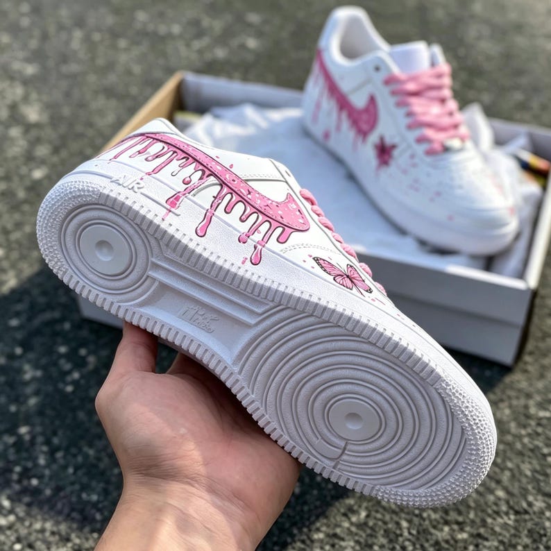 May include: White sneakers with pink accents, including a dripping paint design on the side and a pink butterfly. The sneakers have pink laces and a white sole. The Nike logo is visible on the side.
