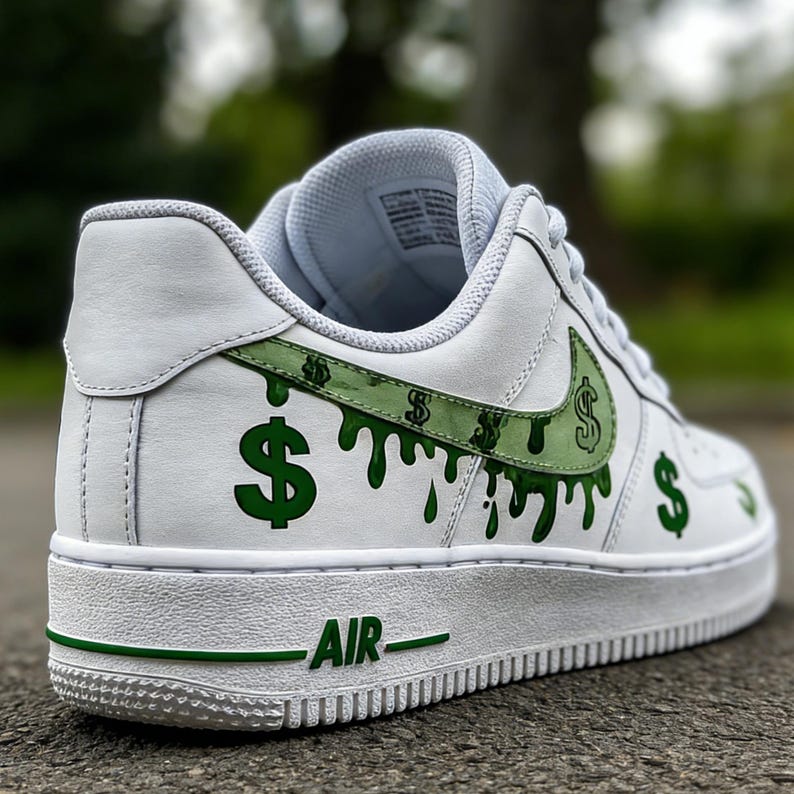 May include: White athletic shoe with green dollar sign accents. The Nike swoosh is green with dollar signs. Green dollar signs drip down the side of the shoe. The word "AIR" is printed in green on the white sole.