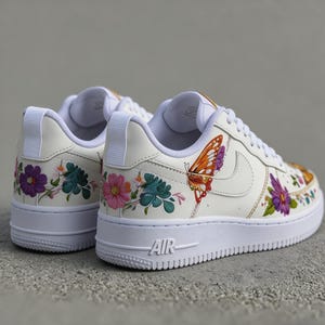 May include: Off-white sneakers with floral and butterfly embroidery. The low-top shoes feature colorful flowers in purple, pink, and teal, and a large orange butterfly. The white soles have the word "AIR" on the side.