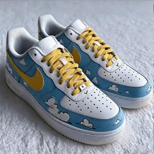 May include: A pair of custom Air Force 1 trainers. The shoes have a white base with blue and yellow accents. The design includes cartoon clouds and yellow laces. The Nike swoosh is yellow.