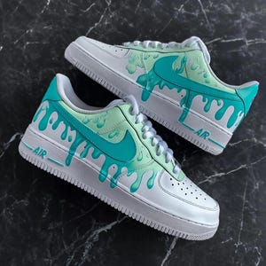 May include: A pair of white sneakers with teal and light green drip design accents. The Nike swoosh is teal, and the word "AIR" is printed on the side. The shoes have white laces and a white sole.