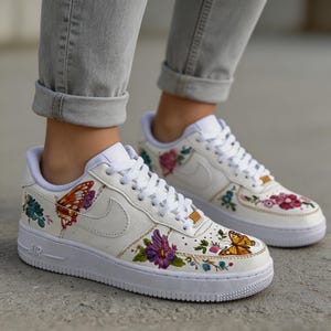 May include: White sneakers with colorful floral and butterfly designs. The shoes feature a low-top design with white laces and soles. The artwork includes a variety of flowers and butterflies in shades of purple, orange, and green.
