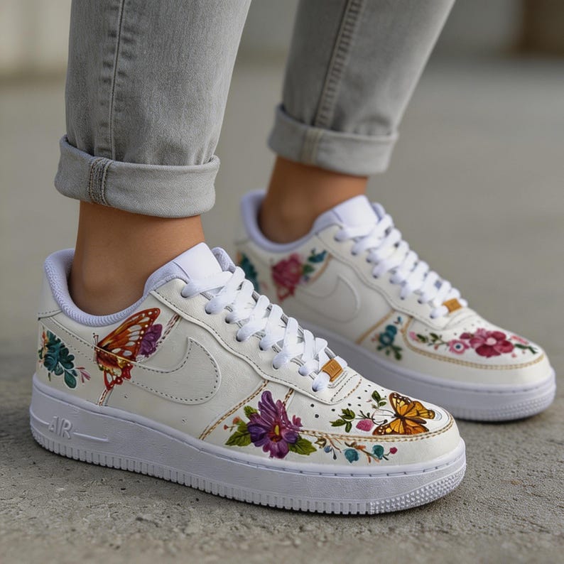 May include: White sneakers with colorful floral and butterfly designs. The shoes feature a low-top design with white laces and soles. The artwork includes a variety of flowers and butterflies in shades of purple, orange, and green.