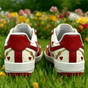 May include: Pair of white sneakers with red accents, including the heel and a Nike logo. The design features red hearts and gold bow and arrow graphics. The shoes are set against a backdrop of green grass and colorful flowers.