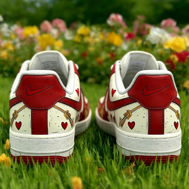 May include: Pair of white sneakers with red accents, including the heel and a Nike logo. The design features red hearts and gold bow and arrow graphics. The shoes are set against a backdrop of green grass and colorful flowers.