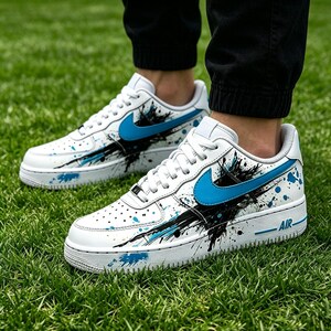 May include: White sneakers with blue and black paint splatter design. The shoes feature a blue Nike swoosh and the word "AIR" in blue on the side. The sneakers are on a green grass background.