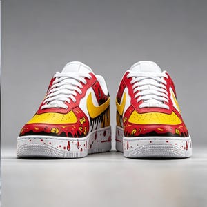 Puede incluir: Un par de zapatillas pintadas a medida. Las zapatillas presentan una base blanca con detalles en rojo y amarillo. El dise&ntilde;o incluye un logotipo de Nike amarillo, gotas rojas y cordones blancos. La entresuela tiene salpicaduras de pintura roja.