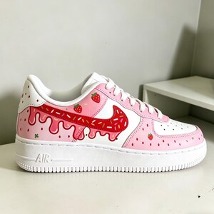 May include: White and pink Air Force 1 sneakers with a strawberry theme. The shoes feature a white sole, pink panels with strawberry details, and a red Nike swoosh with sprinkles. The design includes dripping pink icing.