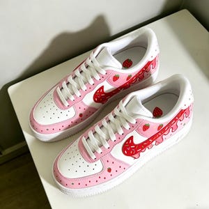 May include: Pair of white and pink sneakers with a strawberry theme. The shoes feature a white base with pink accents, a red swoosh, and strawberry graphics. White laces and soles complete the design.