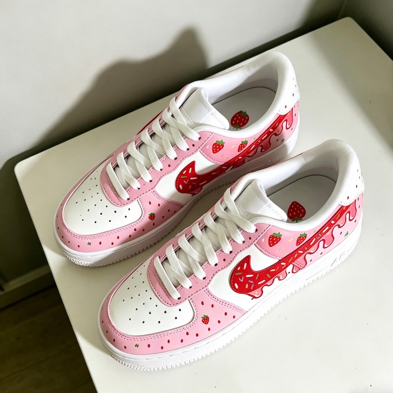 May include: Pair of white and pink sneakers with a strawberry theme. The shoes feature a white base with pink accents, a red swoosh, and strawberry graphics. White laces and soles complete the design.