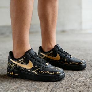 Op de afbeelding: Zwarte en gouden Air Force 1 sneakers met een bliksemontwerp. De sneakers hebben een zwarte basis met gouden accenten op de Nike swoosh en bliksemgraphics. Het woord "AIR" is zichtbaar aan de zijkant.