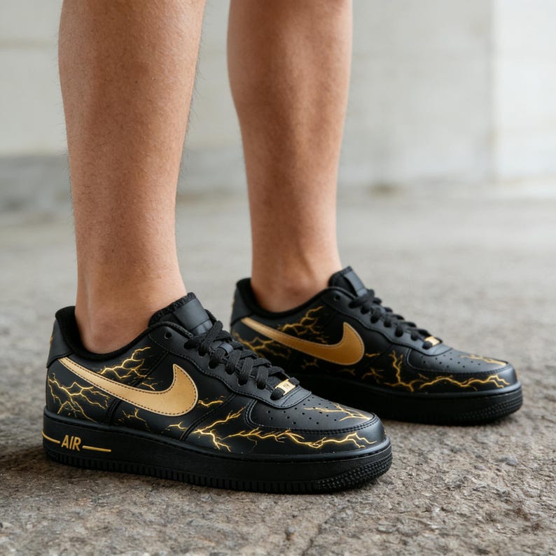 Op de afbeelding: Zwarte en gouden Air Force 1 sneakers met een bliksemontwerp. De sneakers hebben een zwarte basis met gouden accenten op de Nike swoosh en bliksemgraphics. Het woord "AIR" is zichtbaar aan de zijkant.