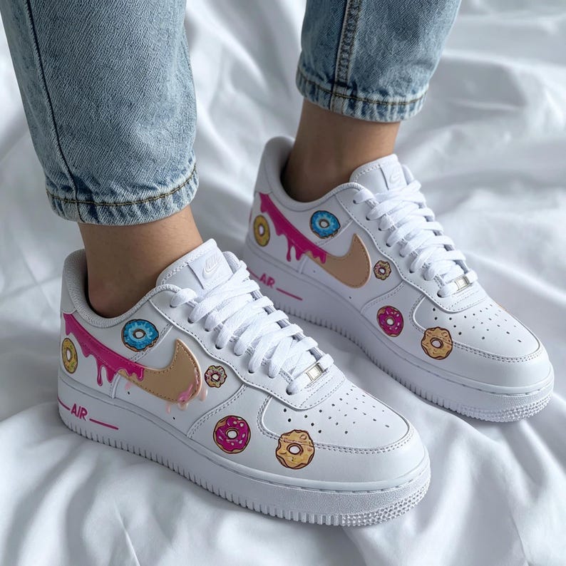 May include: White sneakers with a custom donut design. The shoes feature a pink, blue, and tan donut pattern with a dripping glaze effect. The word "AIR" is printed on the side.