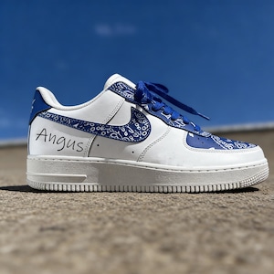 May include: White and blue custom sneaker with a paisley design. The shoe features blue laces and accents, with the name "Angus" written in black on the side. The sneaker is set against a bright blue sky.